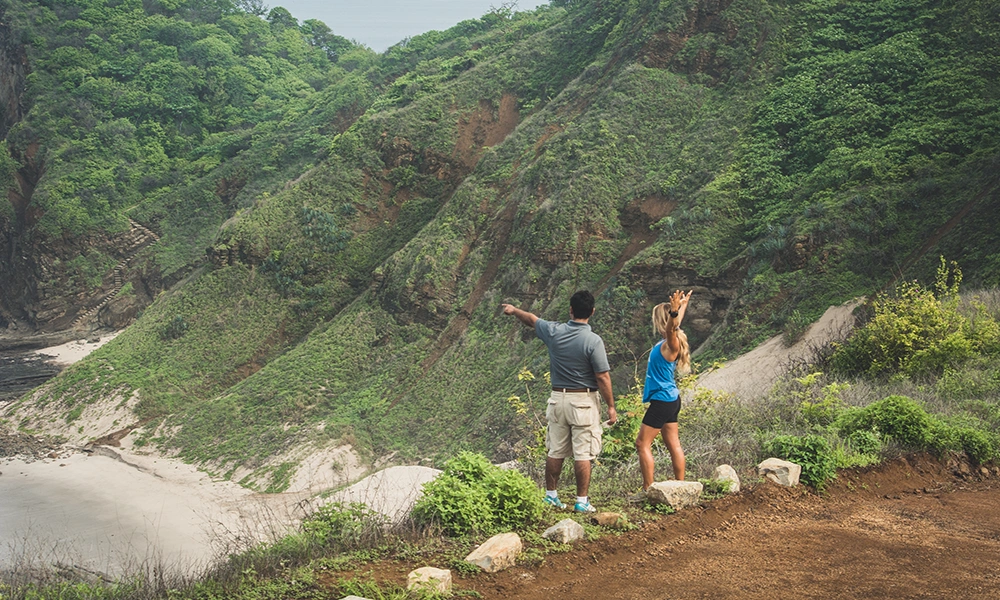 Two hikers enjoying the scenic view on a lush green trail in Nicaragua, perfect for adventure lovers seeking must-visit hiking trails. The trail overlooks a secluded beach surrounded by verdant hills.