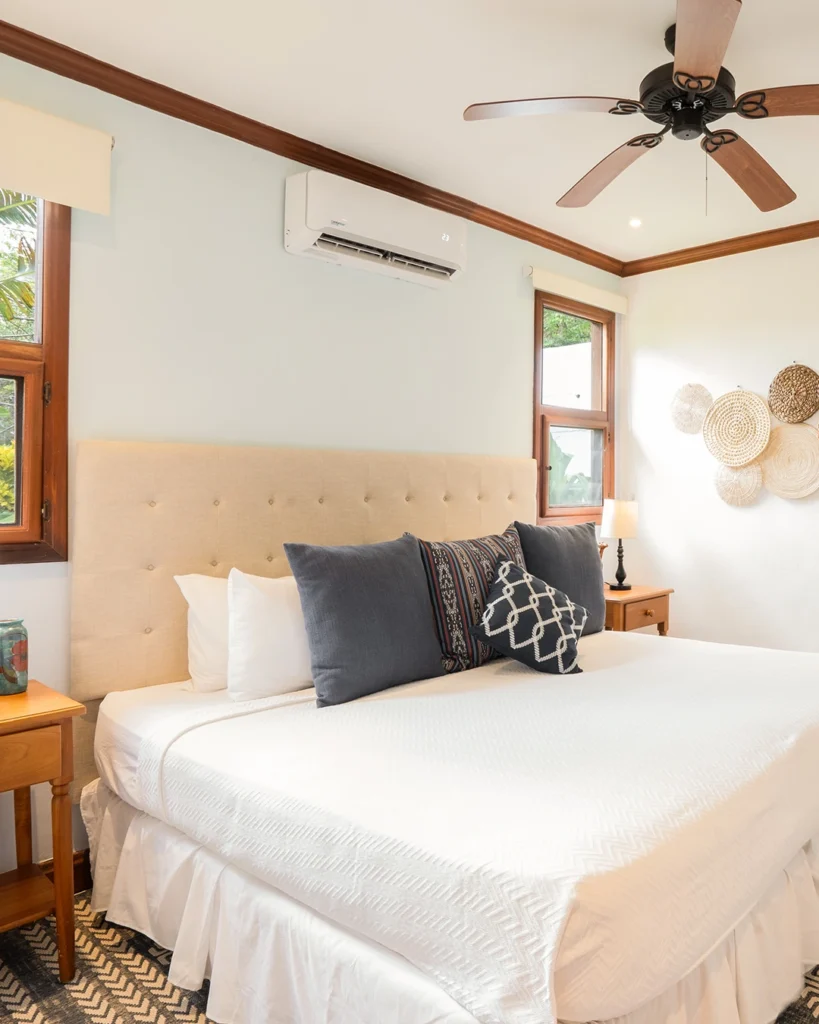 Serene bedroom in Garden View Casita V20 at Rancho Santana, featuring a plush king-sized bed with a tufted headboard, decorative pillows, wooden nightstands with lamps, and a ceiling fan. Natural light flows in through windows, highlighting wall art and creating a peaceful retreat.