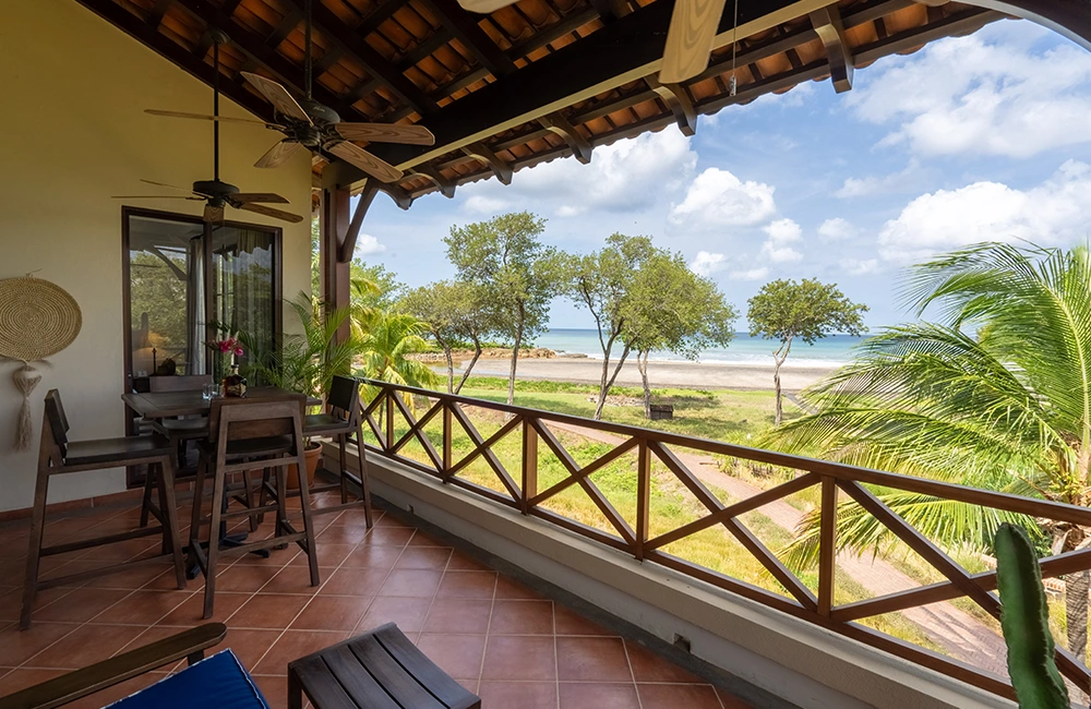 View from the outdoor terrace at Puerta del Mar Villa 4D in Rancho Santana, featuring a wooden dining set under a shaded roof, overlooking palm trees, a green landscape, and the ocean in the distance on a sunny day.