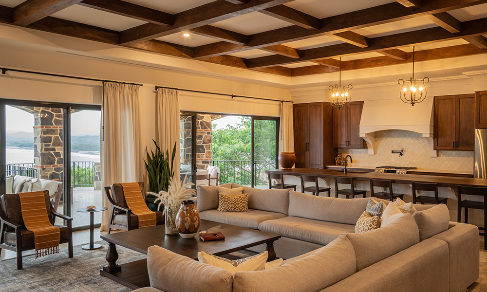 Spacious and elegant living room at Rancho Santana Luxury Collection featuring a large beige sectional sofa, wooden beam ceiling, open kitchen with bar seating, and stunning views of the surrounding nature through sliding glass doors.