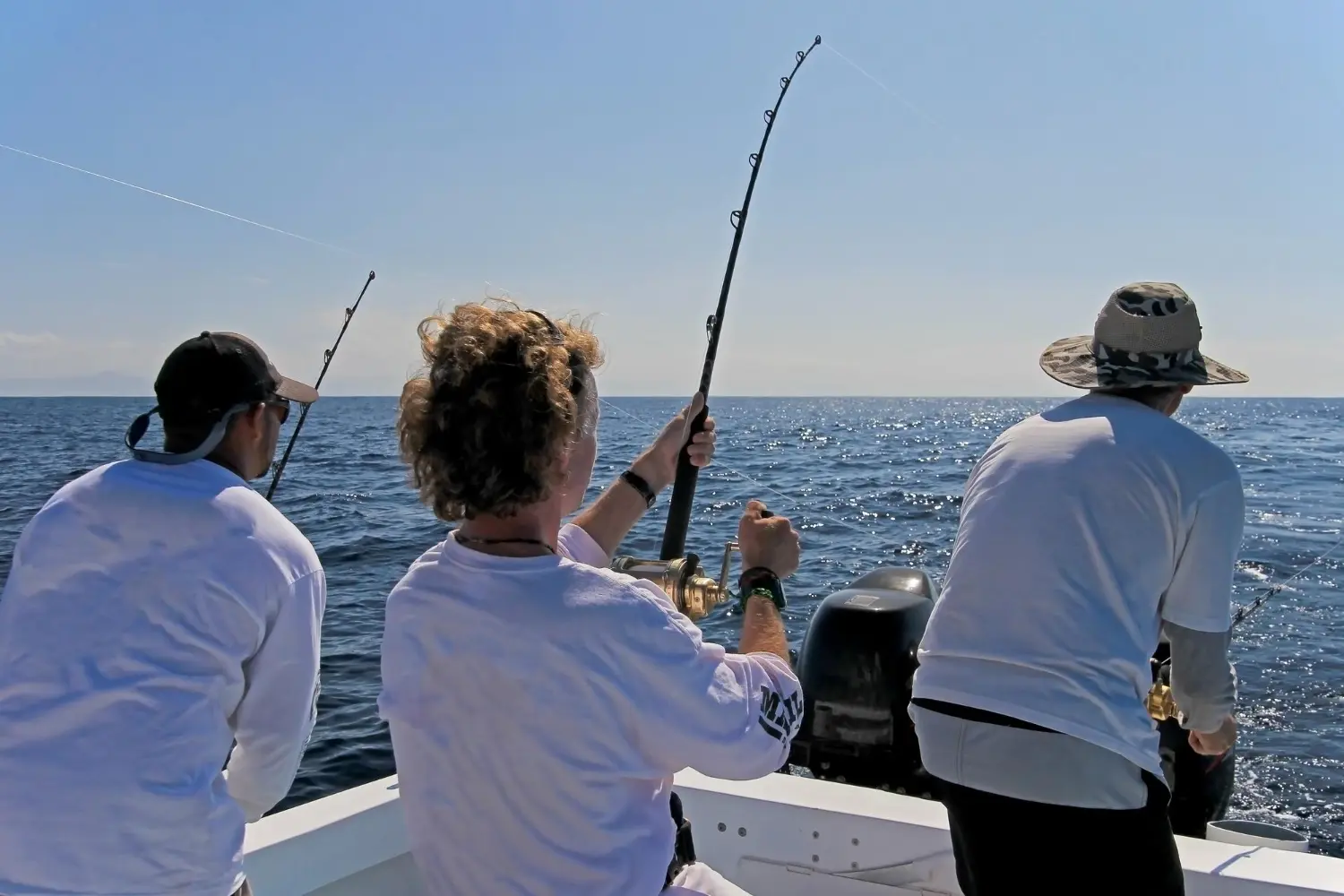Three anglers deep sea fishing in the Emerald Coast of Nicaragua, reeling in a catch from a boat under a clear blue sky.
